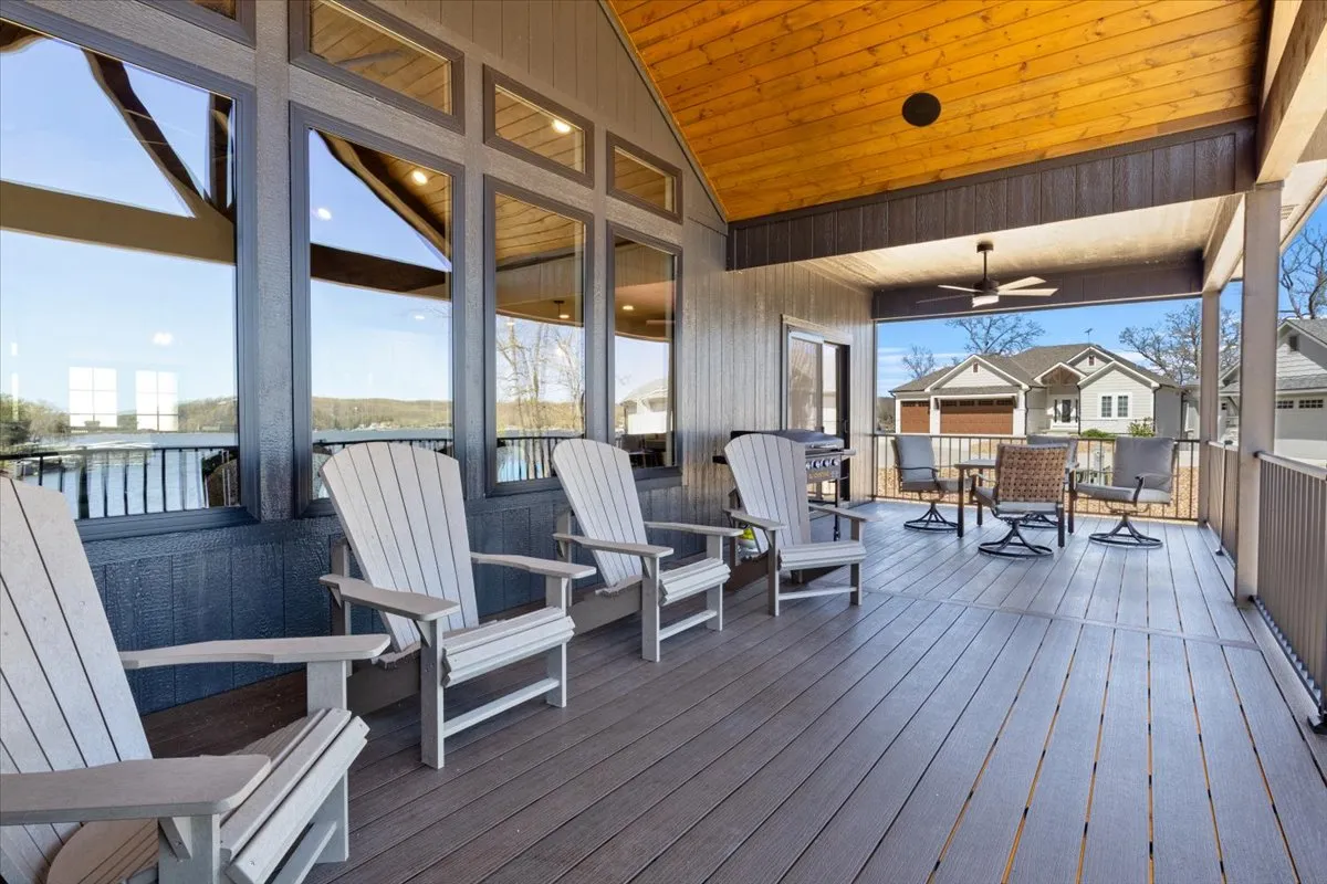 The image showcases an inviting outdoor seating area featuring a row of Adirondack chairs arranged under a wooden ceiling, set against the backdrop of expansive windows that reflect the tranquil lakeside surroundings. The space is clearly designed for relaxation, conversation, and taking in the stunning sunset views over the water.