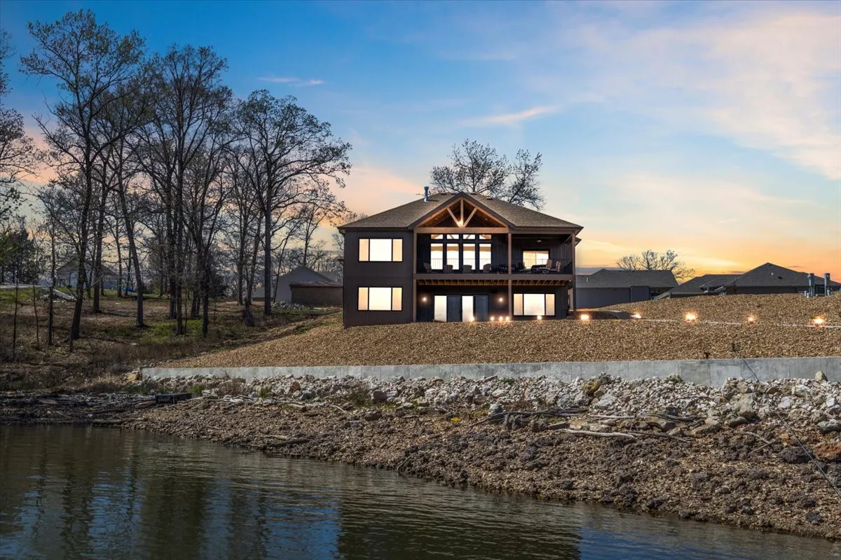 This image captures the modern architecture of Hawk's Cove, featuring large windows and wooden beams that open up to expansive lake views. The scene evokes a sense of connection to the water, with visible areas designed for relaxing by the lake, entertaining guests, and enjoying sunset views.