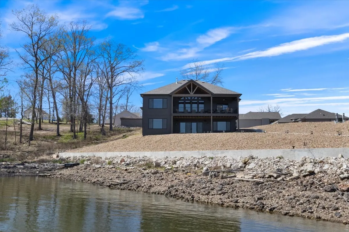 This exterior image showcases a modern lakefront house surrounded by trees, with direct access to the water. The view captures the peaceful setting and hints at activities like boating, fishing, and nature walks, while highlighting the scenic beauty of the Lake of the Ozarks.