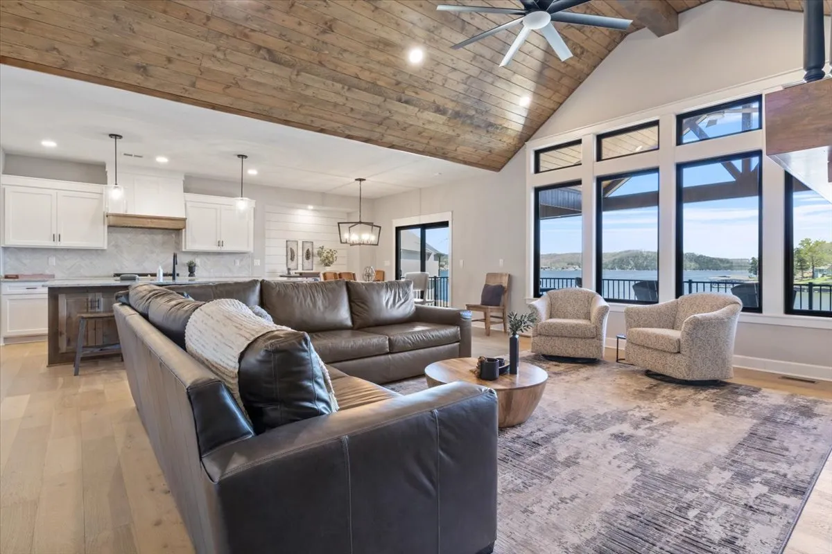 A spacious main living room featuring a large leather sofa, a wooden ceiling, and expansive windows that reveal beautiful sunset views over the lake. The coffee table is set for casual gatherings, and the space is designed for both relaxation and entertaining.